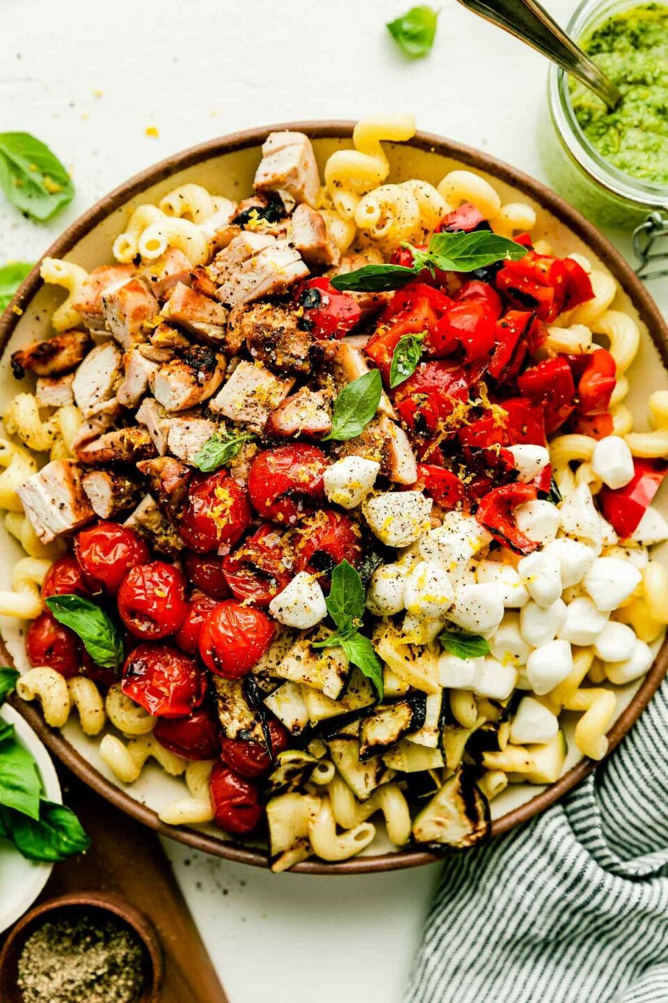 An overhead shot of a large bowl of prepared pasta salad ingredients, sitting alongside a wooden cutting board, bowl of basil leaves, striped dish towel, jar of pesto, and bowl of pepper atop a white surface.