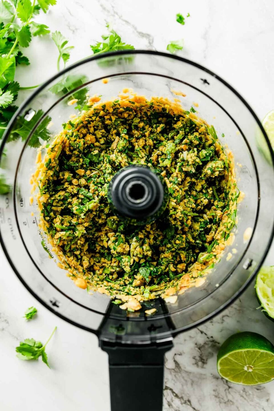 An overhead shot of an uncovered food processor with chopped hummus ingredients atop a white marbled surface.