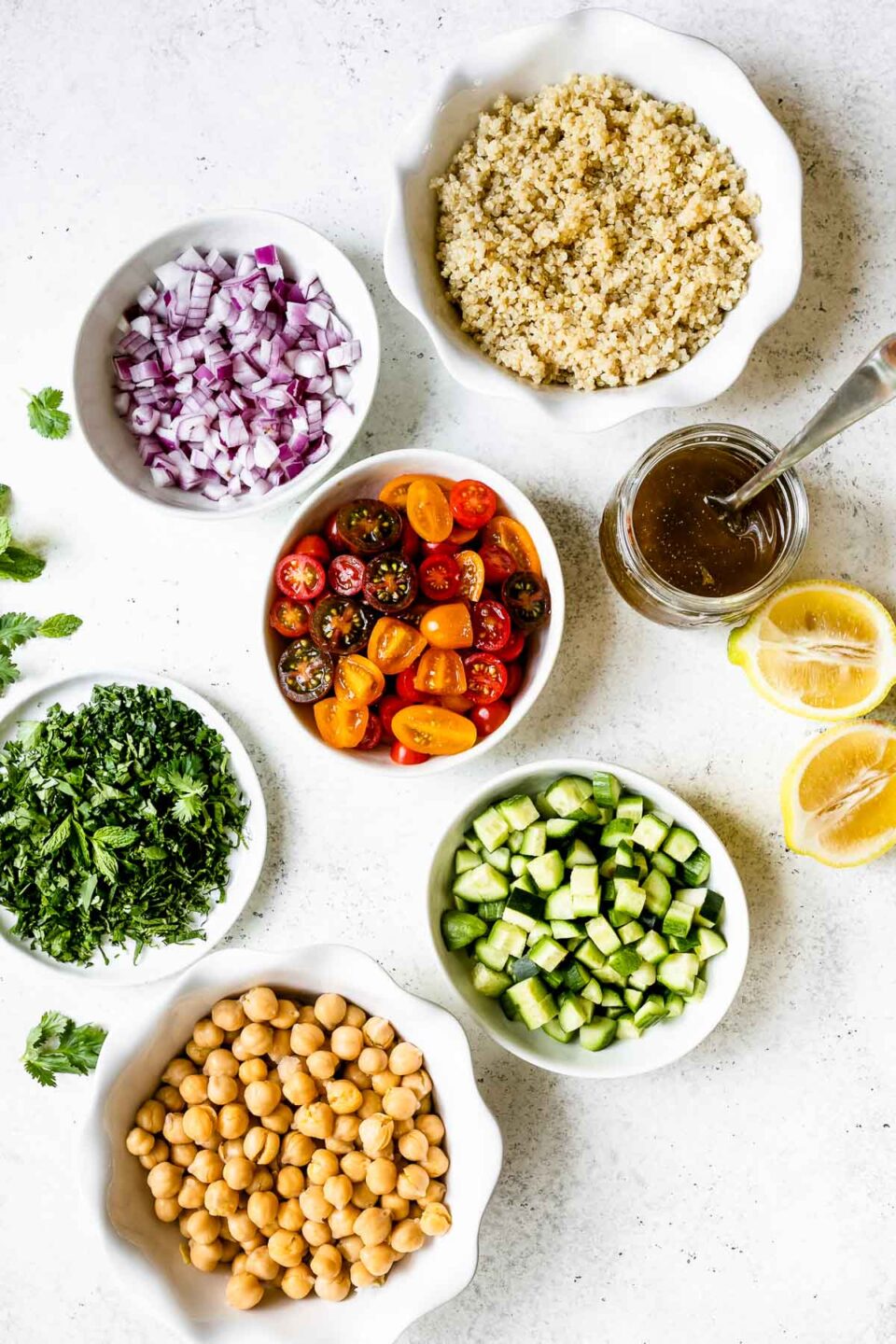 Quinoa salad ingredients in individual small bowls, arranged on a white surface - red onion, quinoa, tomatoes, salad dressing, lemon, cucumber, chickpeas & fresh herbs.