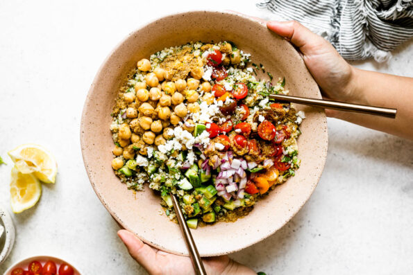 Falafel-ish Quinoa Salad in a large, pink serving bowl. A woman's hands hold the bowl up off of a white surface. Surrounding the salad are a linen napkin, & some lemon wedges.