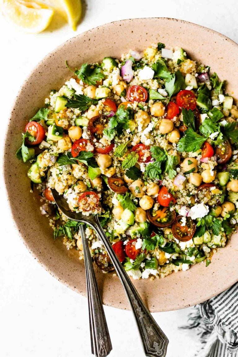Close up of Falafel-ish Quinoa Salad shown in a large pink ceramic serving bowl. The salad is topped with lots of fresh parsley leaves. Two vintage spoons are nestled in the left side of the bowl. The bowl sits atop a white surface, with a gray & white striped linen nestled next to it.