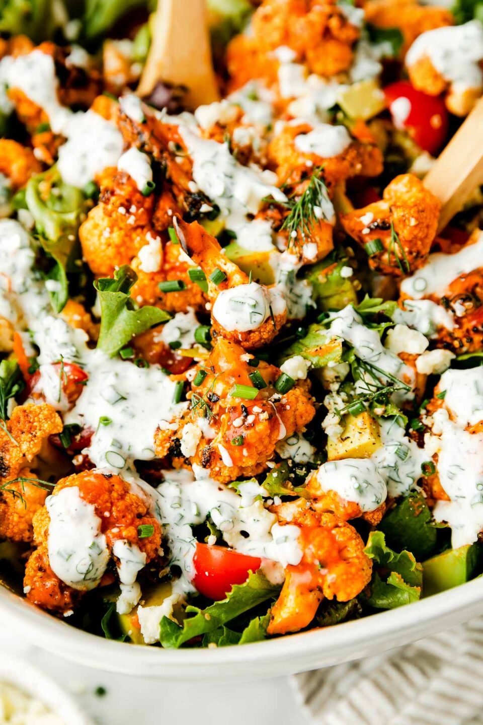 A close-up shot of grilled buffalo cauliflower salad with herbed yogurt ranch drizzled over the top in a white bowl atop a white surface.