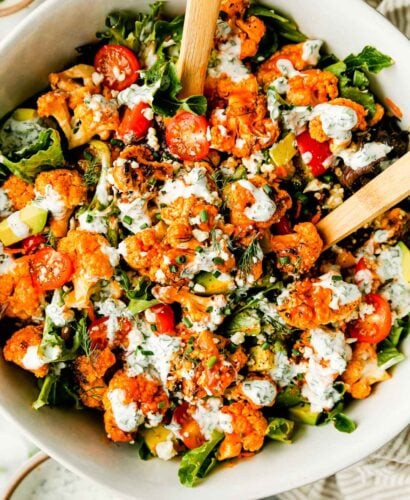 An overhead shot of a large bowl of grilled buffalo cauliflower salad with wooden serving spoons atop a striped dish towel on a white marbled surface. A bowl of herbed yogurt ranch and small bowls of herbs sit beside it.