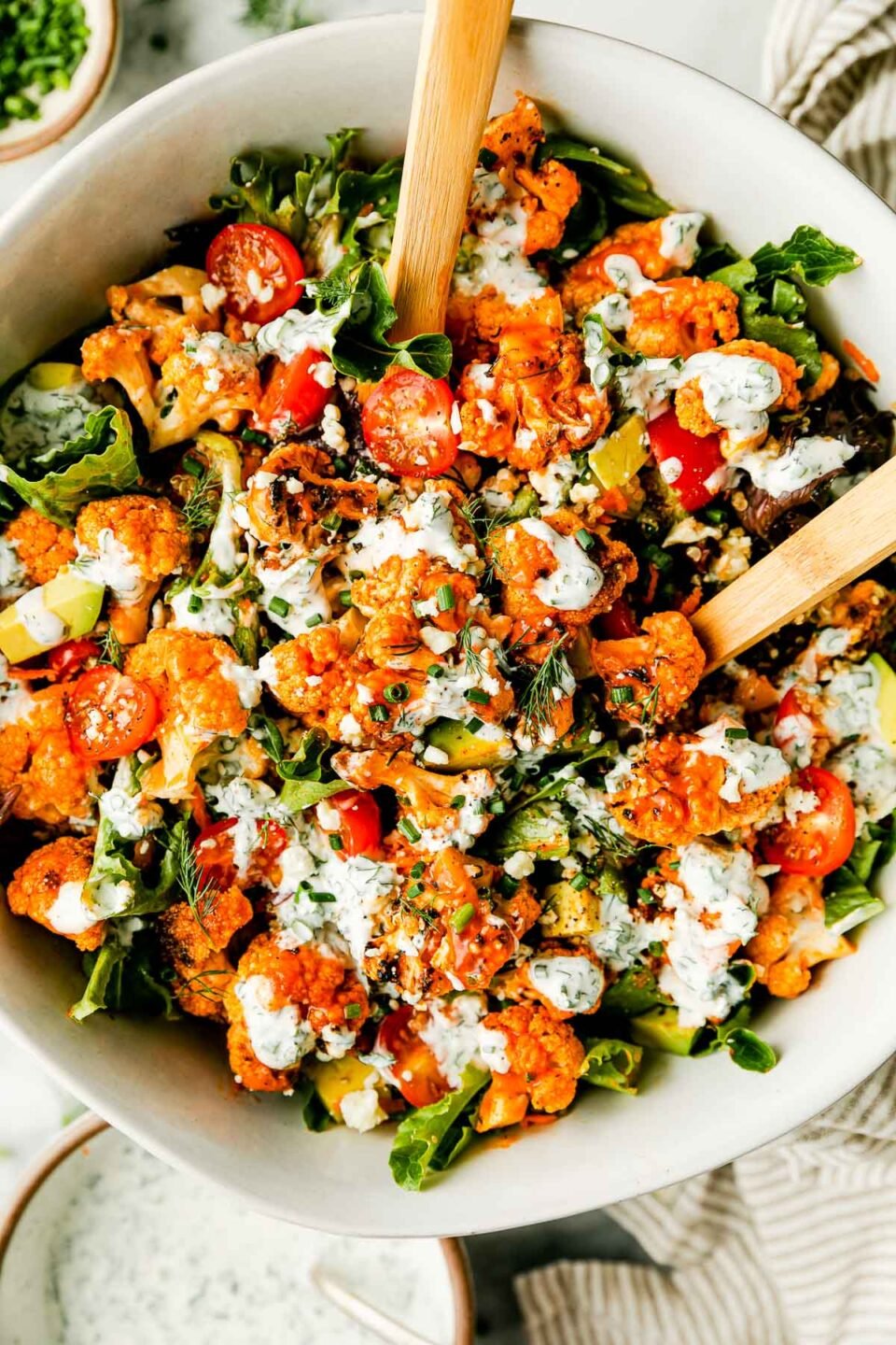 An overhead shot of a large bowl of grilled buffalo cauliflower salad with wooden serving spoons atop a striped dish towel on a white marbled surface. A bowl of herbed yogurt ranch and small bowls of herbs sit beside it.