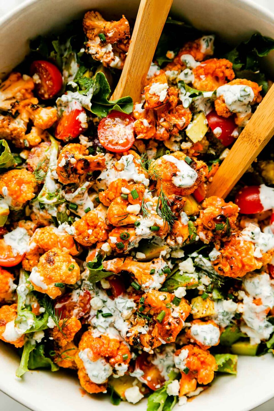An overhead shot of a large white bowl of grilled buffalo cauliflower salad with wooden serving spoons.