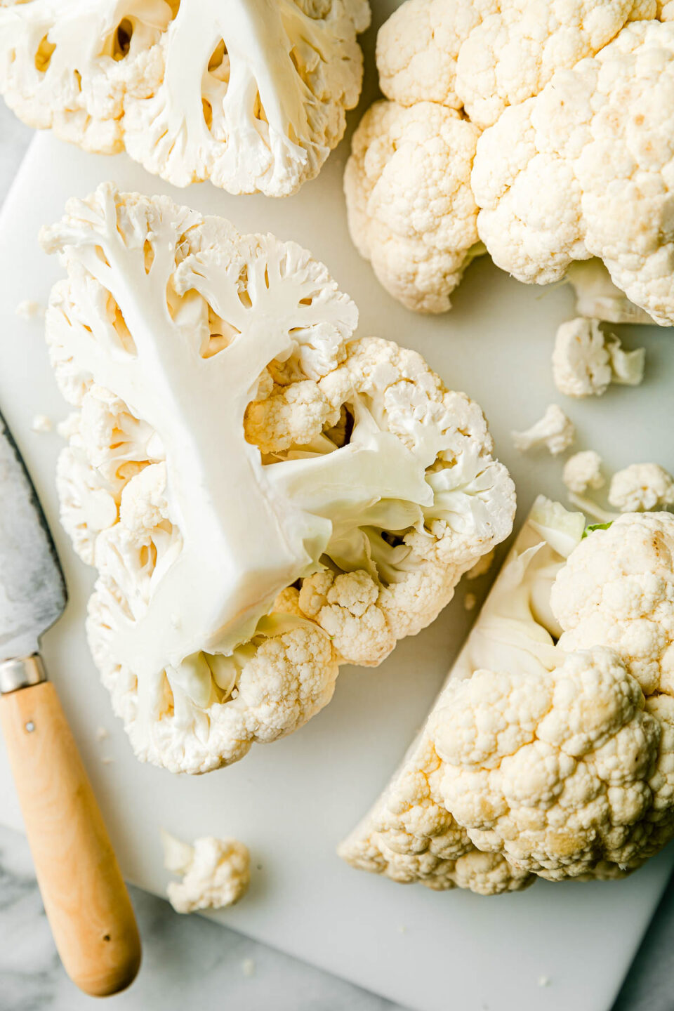 An overhead shot of cauliflower with a chef's knife on a white cutting board atop a white surface.
