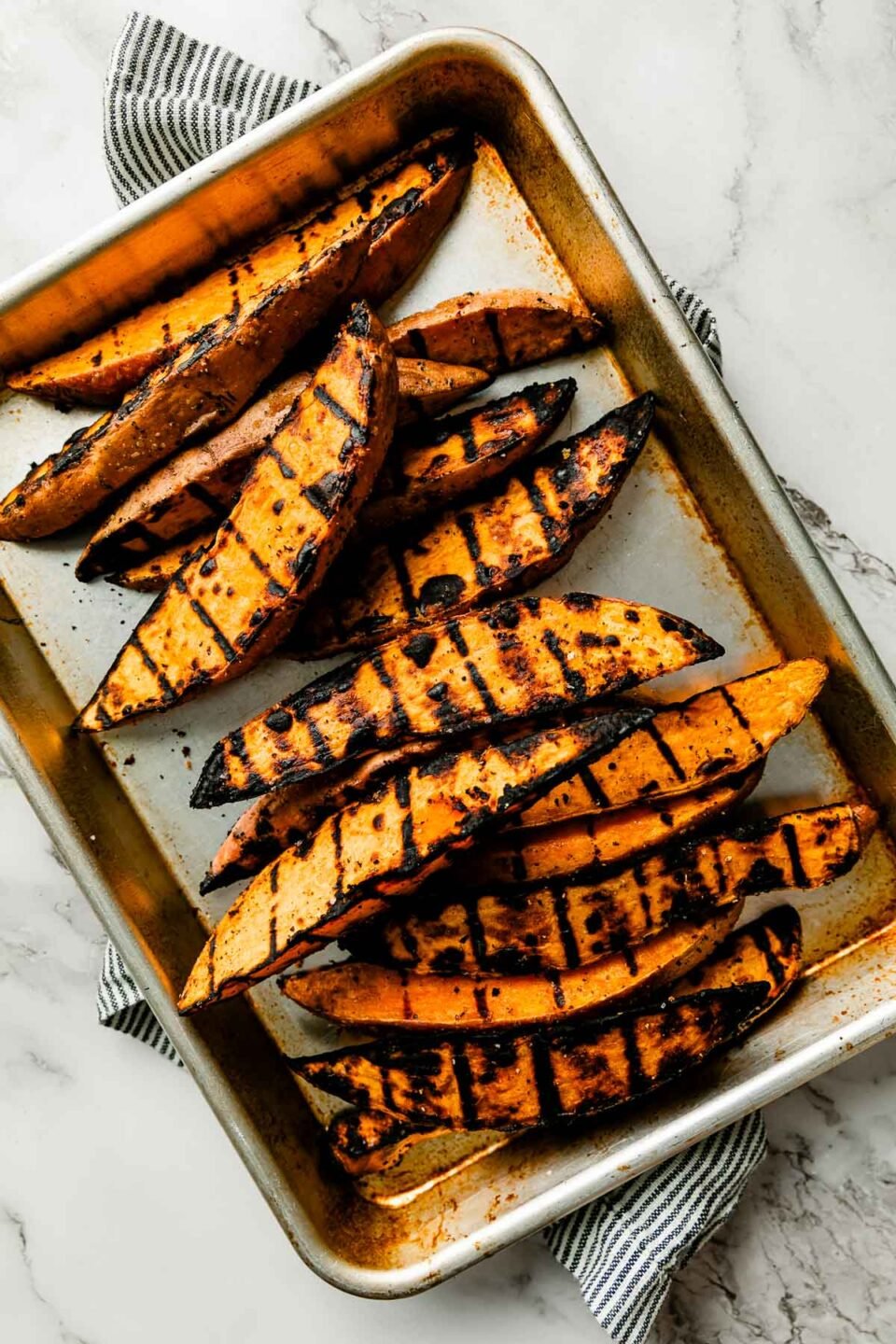 Grilled sweet potato wedges on a metal quarter baking sheet atop a striped linen napkin on a creamy white marble surface.