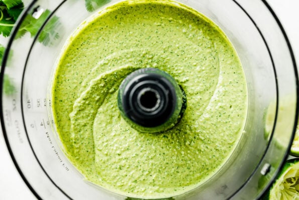A close-up overhead shot of an uncovered food processor with finished smooth cilantro jalapeno hummus, resting atop a white marbled surface alongside squeezed limes and cilantro leaves.