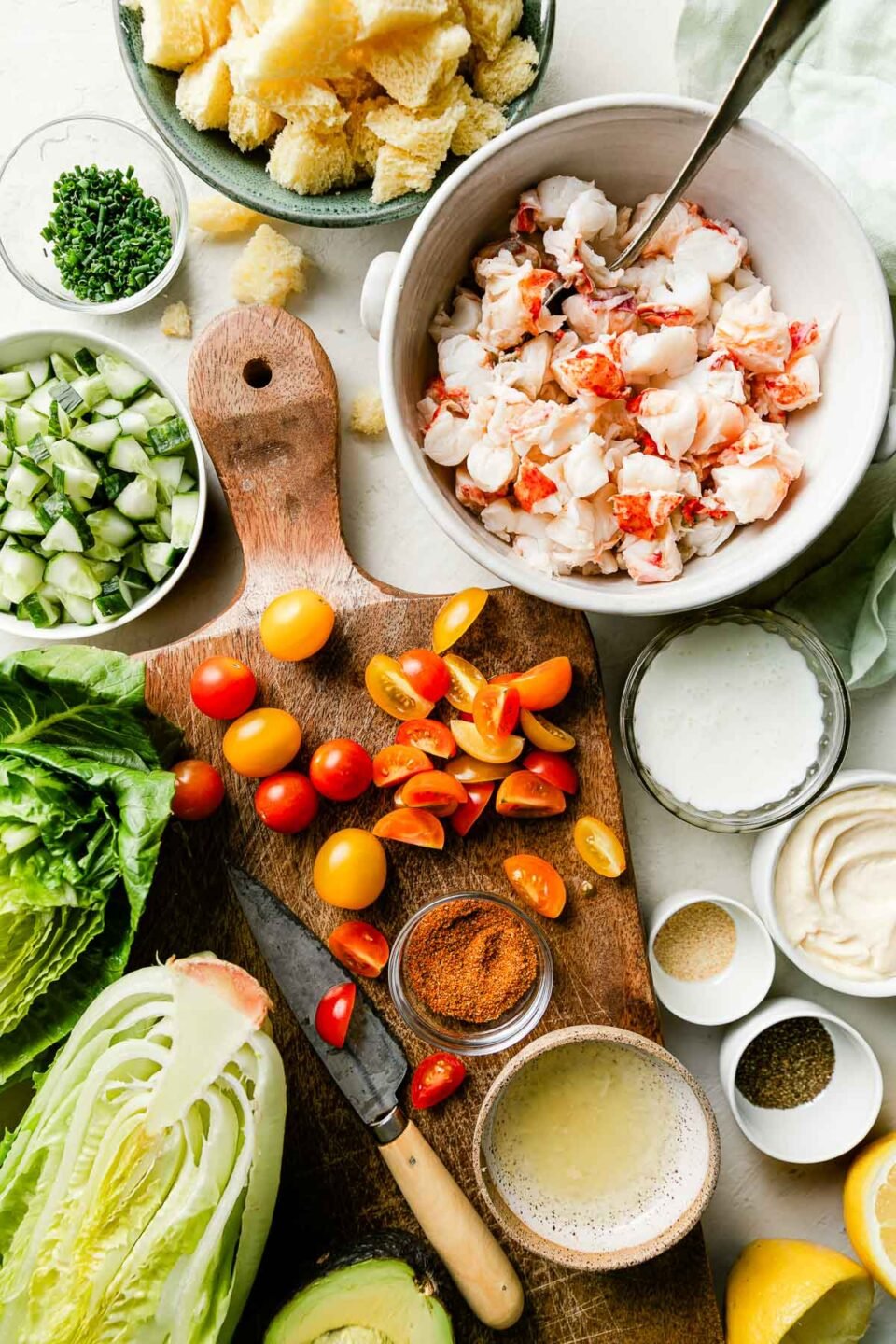 Lobster salad ingredients are arranged on a wooden cutting board and white surface: lettuce, sliced cherry tomatoes, chopped cucumbers, chunks of brioche bread, chopped lobster, and dressing ingredients.