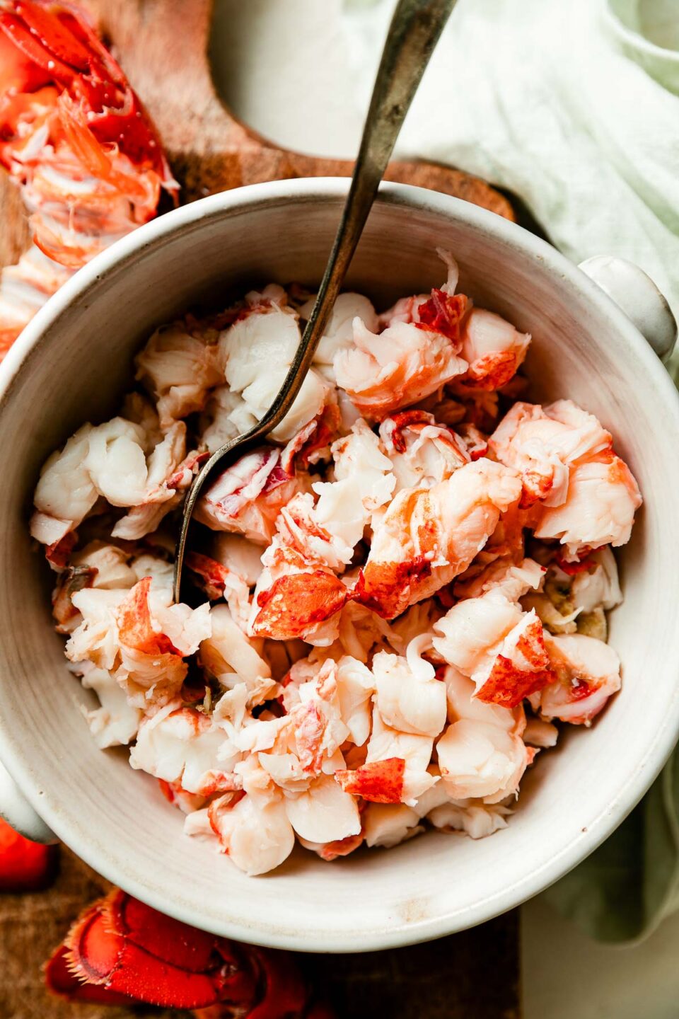 An overhead shot of a white bowl of chopped cooked lobster atop a wooden cutting board.