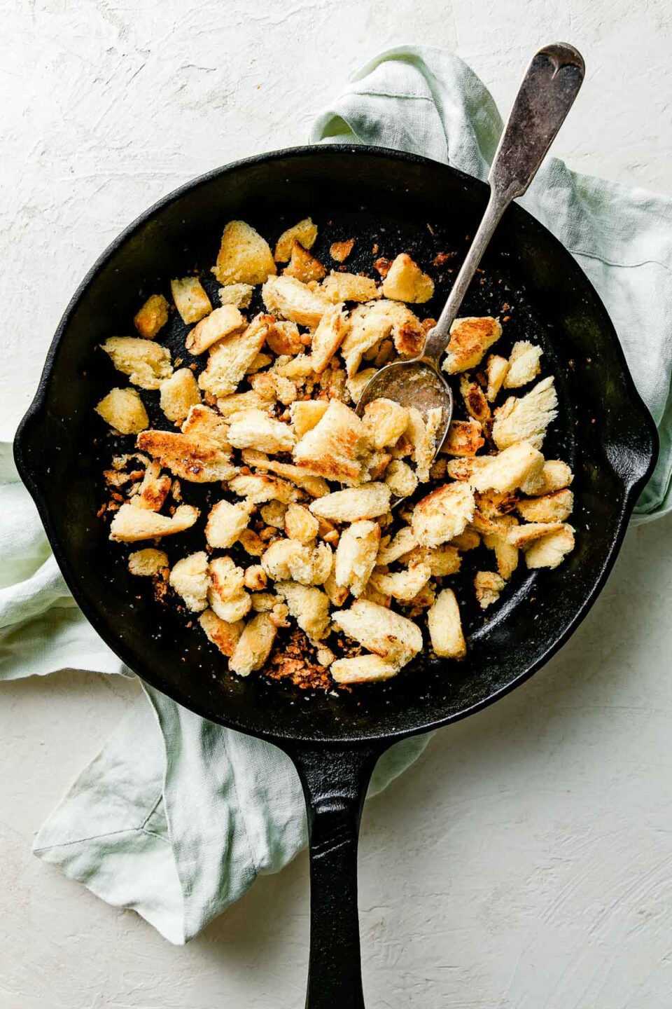 An overhead shot of a black skillet with browned brioche breadcrumbs and a spoon resting in it. The skillet sits atop a light green cloth atop a textured white surface.