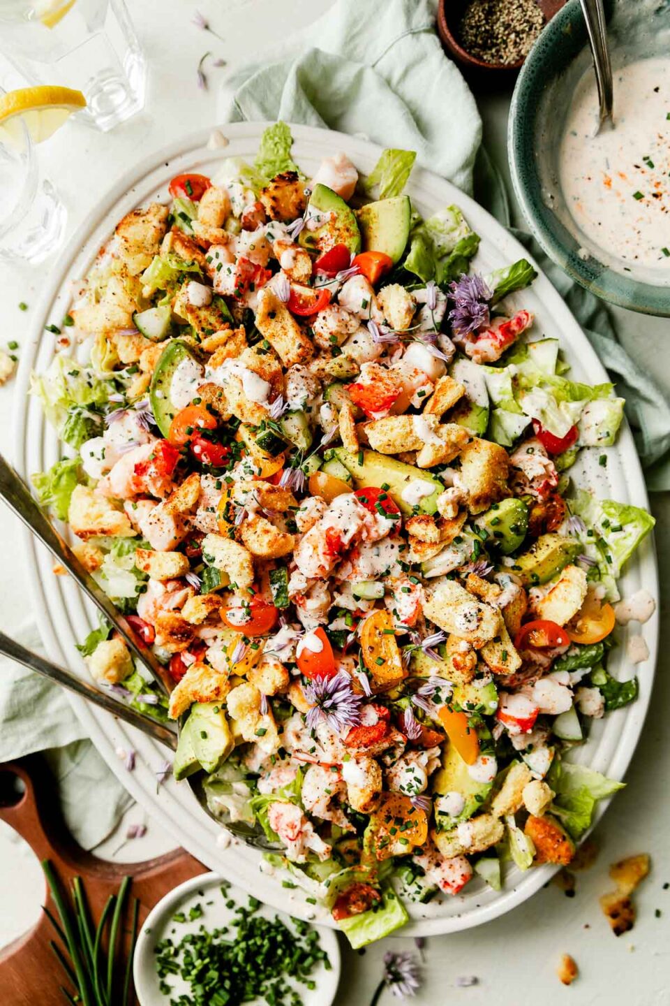 An overhead shot of a white oval platter of lobster salad atop a green cloth on a white textured surface. A small green bowl of dressing, two glasses of water and small dishes of chives and pepper sit alongside the platter.