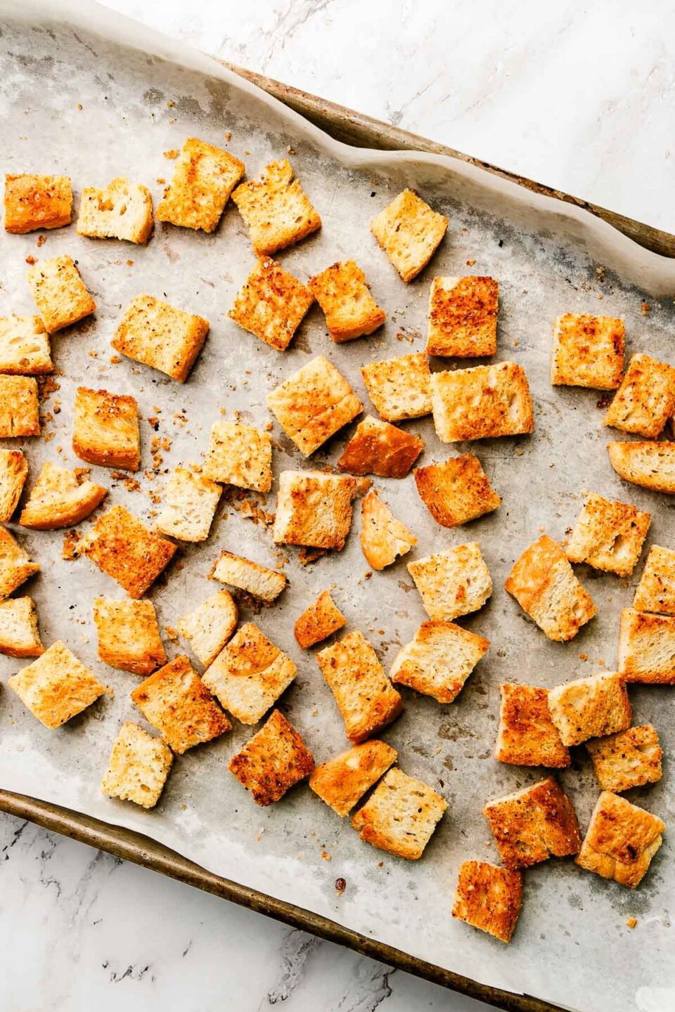 An overhead shot of sourdough croutons on a parchment paper-covered sheet pan, sitting atop a white marbled surface.