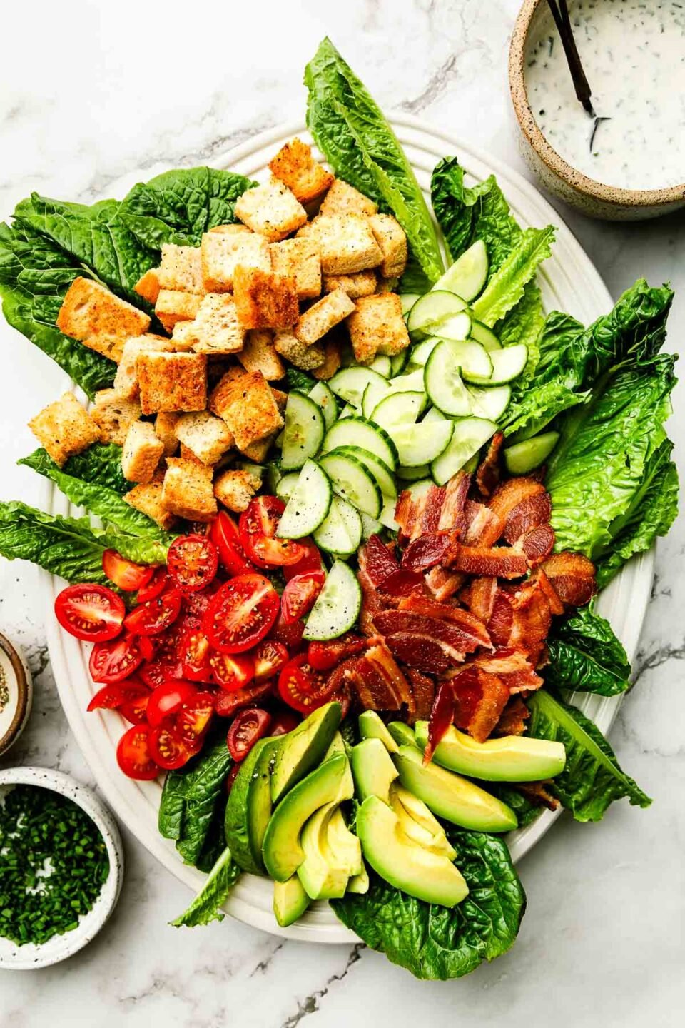 An overhead shot of BLT Salad components on a white oval platter: Lettuce, cucumbers, cherry tomatoes, bacon, avocado, and croutons.