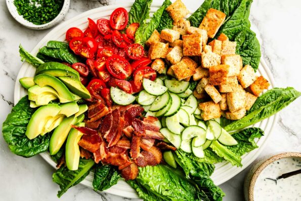 An overhead shot of BLT Salad components on a white oval platter: Lettuce, cucumbers, cherry tomatoes, bacon, avocado, and croutons.