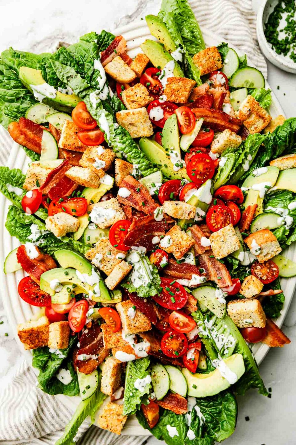 An overhead shot of assembled and dressed BLT salad on a white oval platter atop a white marbled surface.