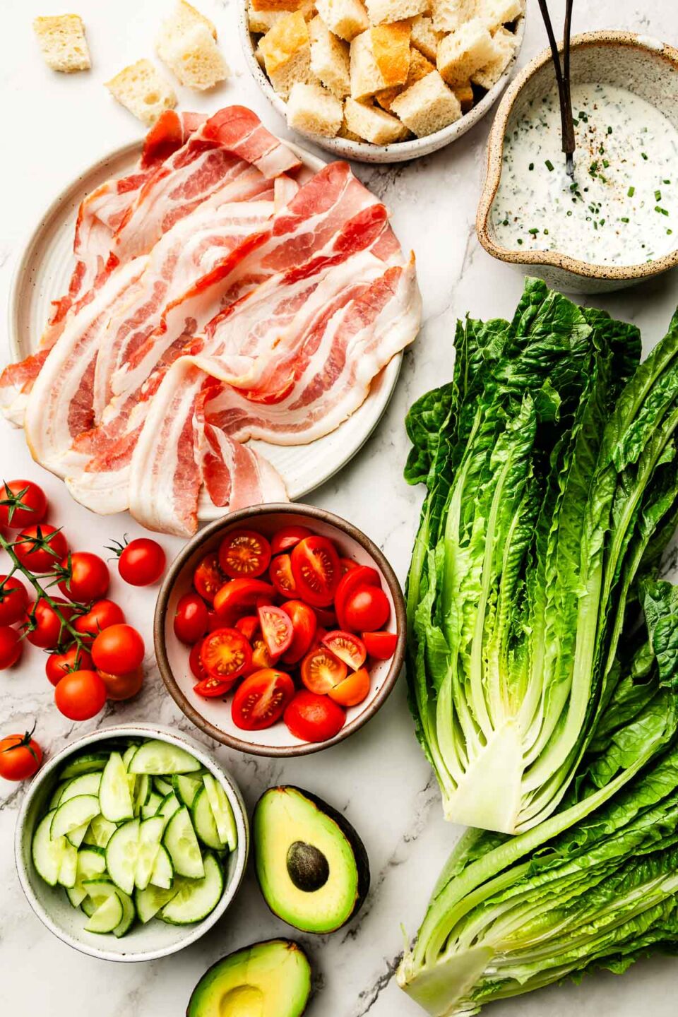 An overhead shot of ingredients on a white marbled surface: lettuce, sliced cherry tomatoes, raw bacon, halved avocado, cubed sourdough, sliced cucumbers and a bowl of dressing.