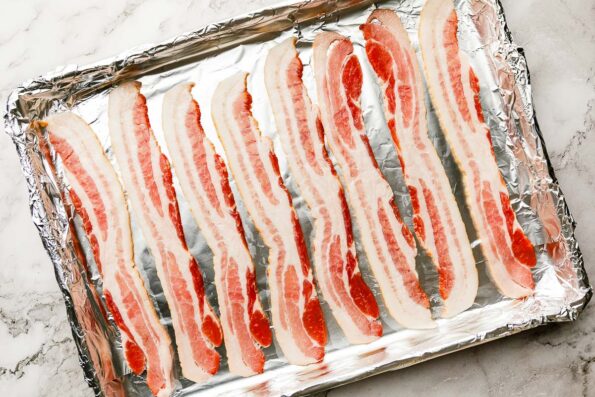 An overhead shot of raw bacon slices on a foil-lined sheet pan, sitting atop a white marbled surface.