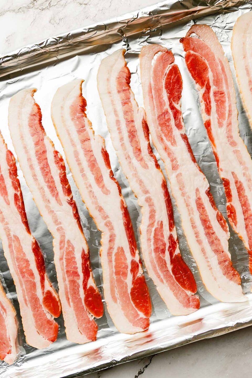An overhead shot of raw bacon slices on a foil-lined sheet pan, sitting atop a white marbled surface.