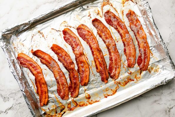 An overhead shot of cooked bacon slices on a foil-lined sheet pan, sitting atop a white marbled surface.