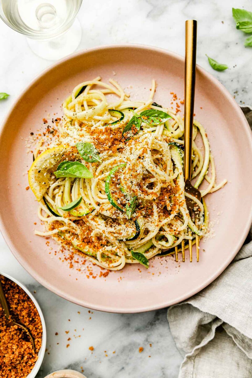 An overhead shot of lemon zucchini pasta on a pink plate atop a cloth napkin on a white marbled surface. A gold fork sits on the plate, and a dish of toasted breadcrumbs rests beside it.