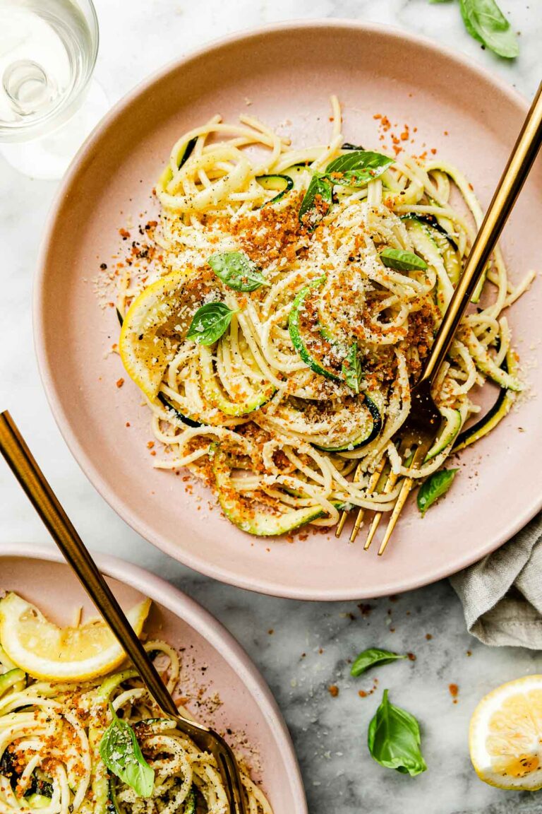 An overhead shot of lemon zucchini pasta on a pink plate atop a white surface. A second plate of pasta sits beside it.