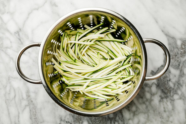 An overhead shot of julienned zucchini in a colander atop a white and grey marbled surface.
