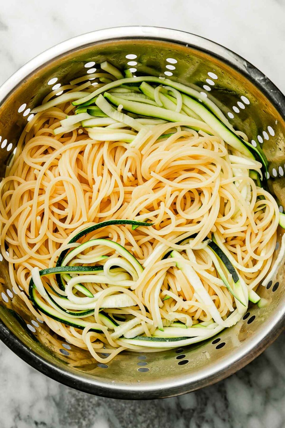 An overhead shot of julienned zucchini and cooked spaghetti in a colander atop a white and grey marbled surface.