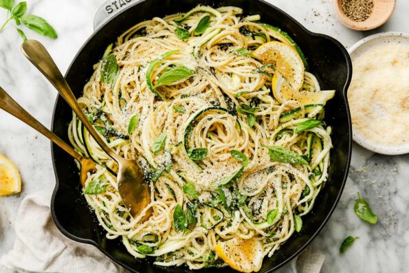 An overhead shot of lemon zucchini pasta garnished with basil and lemon wedges in a black skillet atop a white marbled surface. The skillet is surrounded by a dish of parmesan, black pepper, a cloth napkin, basil leaves and lemon slices.