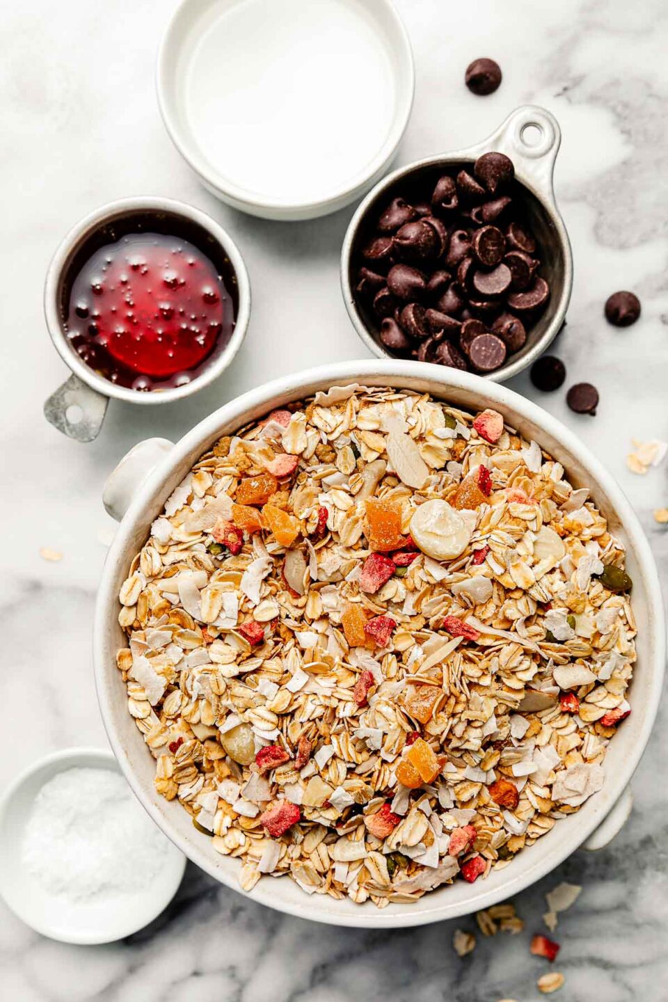 Ingredients are displayed in white bowls atop a white and grey marbled surface: dry muesli, salt, melted coconut oil, chocolate chips, and maple syrup.