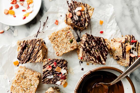An overhead shot of sliced muesli bars on parchment paper atop a white marbled surface, some drizzled with chocolate and some plain. A bowl of chocolate sauce and a plate of muesli sit beside them.