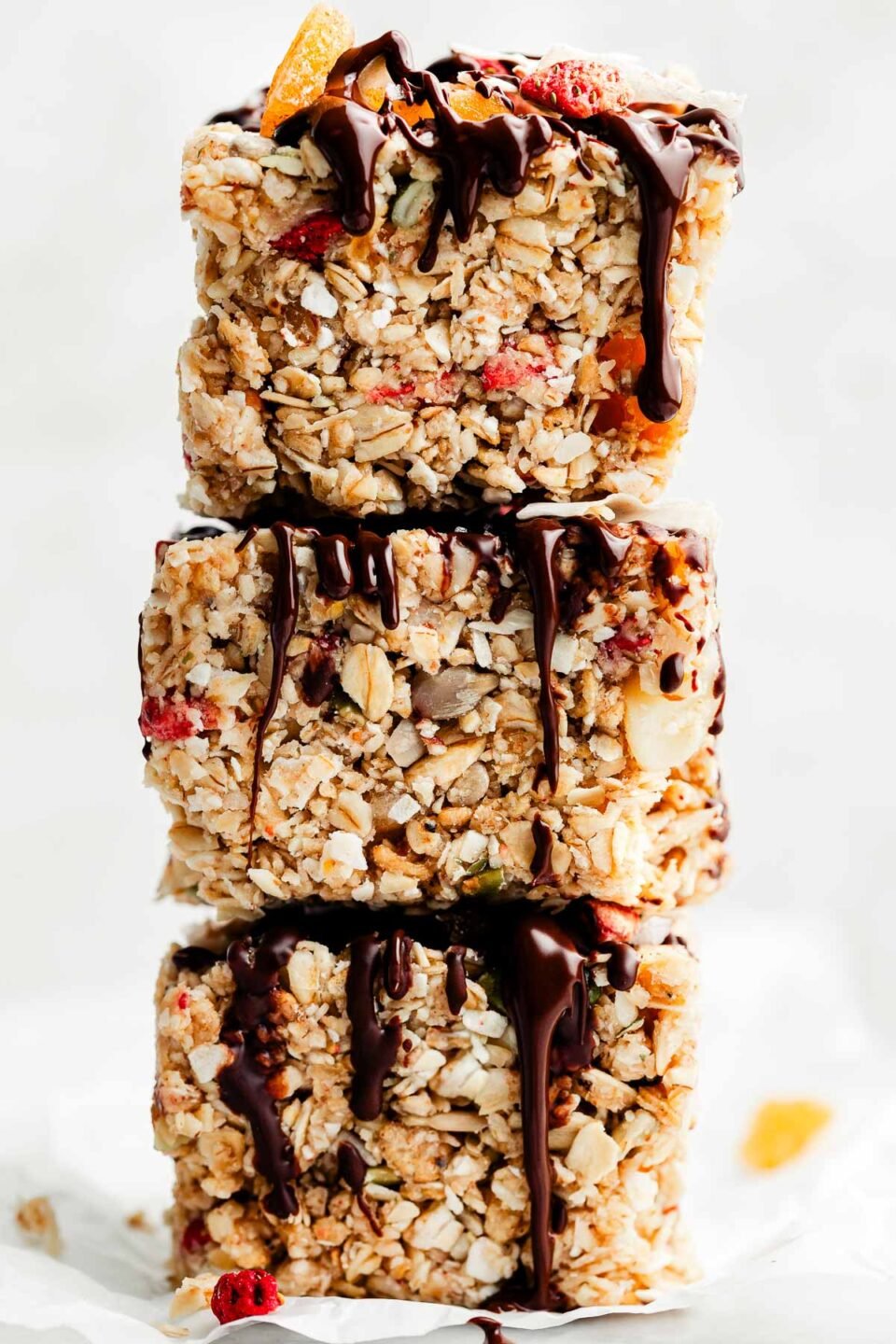A side shot of a stack of three chocolate-drizzled muesli bars sitting on a piece of white parchment paper in front of a white background.