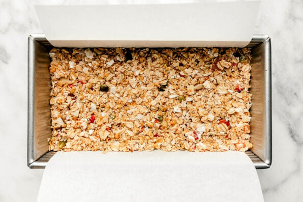 An overhead shot of a silver loaf pan with parchment paper and prepared muesli bars, sitting atop a white and grey marbled surface.