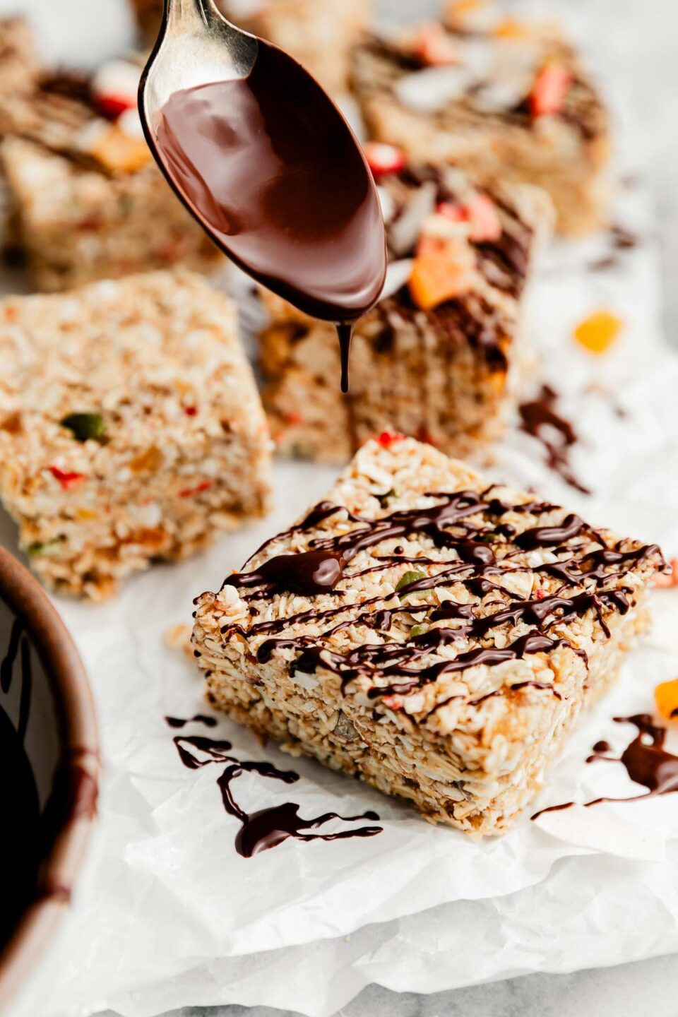 A side shot of a sliced muesli bar on parchment paper being drizzled with chocolate sauce from a spoon. Chocolate drizzled and plain muesli bars sit in the background.