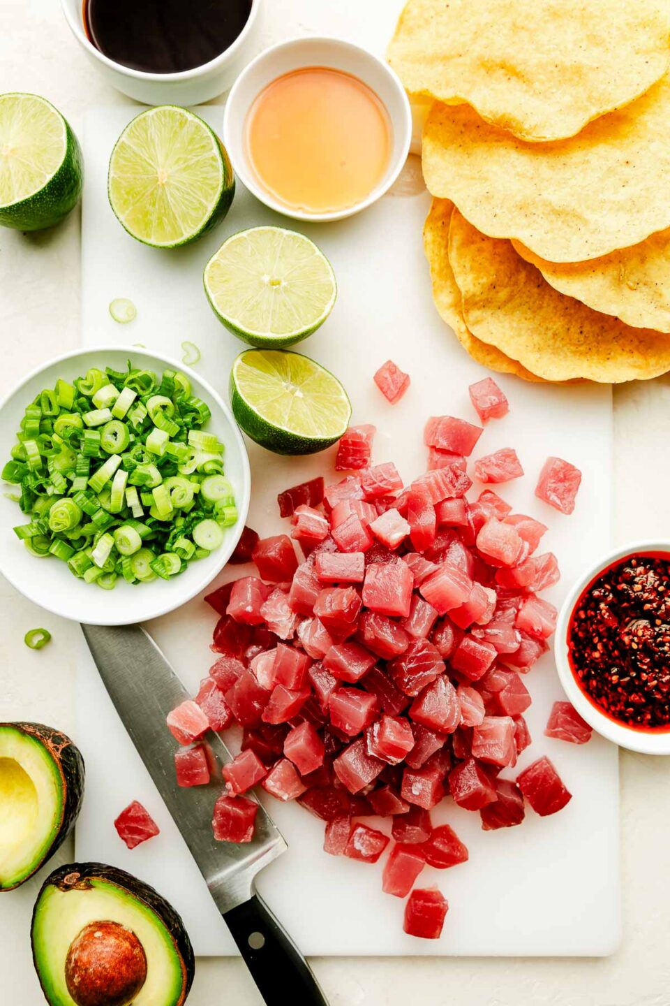 An overhead shot of ingredients arranged on a white cutting board atop a white surface: cubed ahi tuna, tostada shells, halved limes, halved avocado, green onions, soy sauce, sesame oil, and chili crisp.