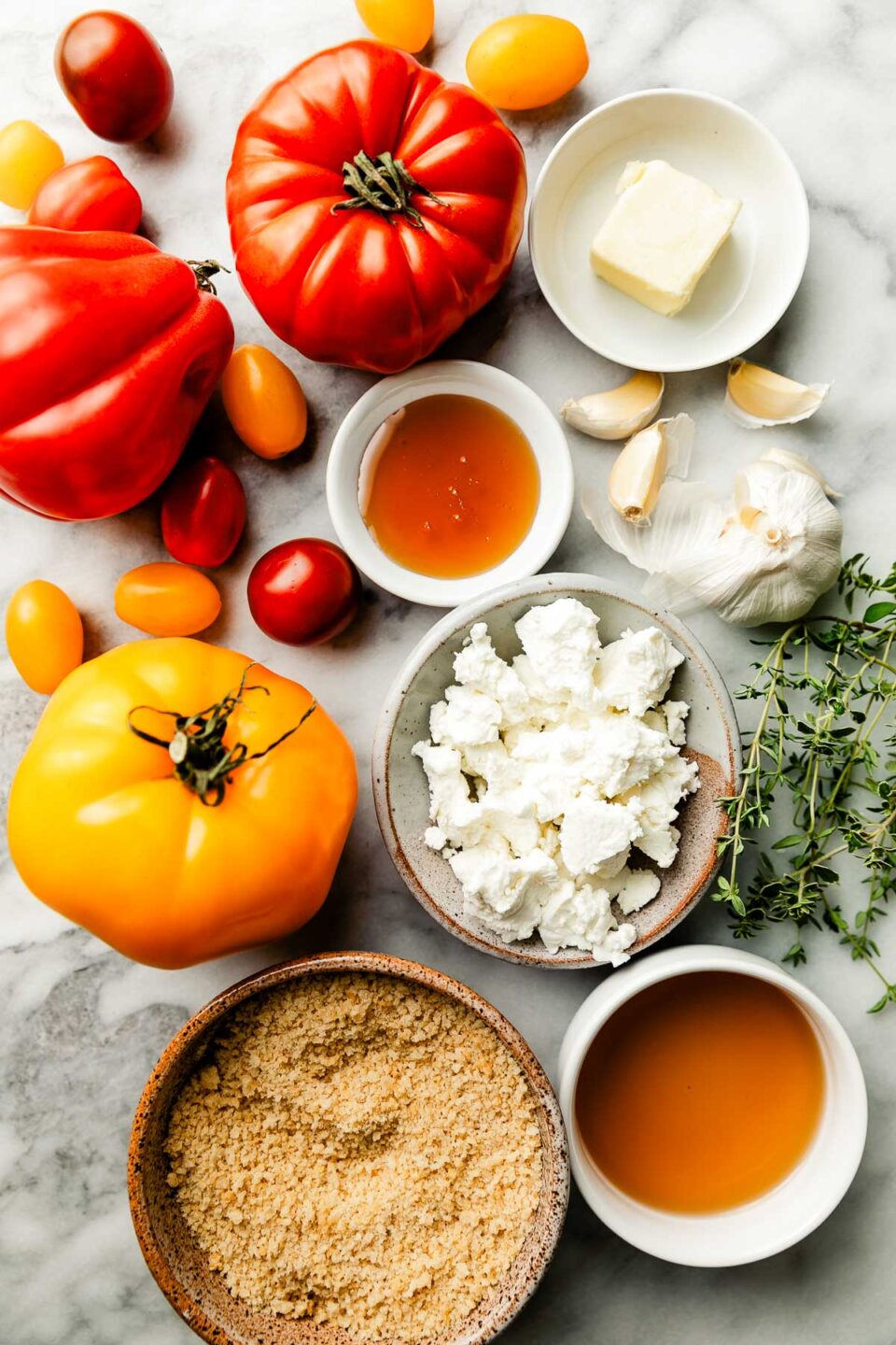 Ingredients displayed on a white and grey marbled surface: bowls of crumbled goat cheese, honey, butter, white balsamic vinegar, salt, thyme sprigs and heirloom and cherry tomatoes.