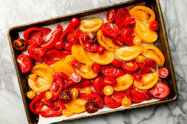An overhead shot of sliced tomatoes seasoned with salt on a cookie sheet atop a white and grey marbled surface.