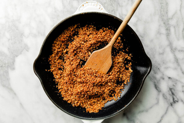 An overhead shot of toasted garlic breadcrumbs with a wooden spoon in a black skillet, sitting atop a white and grey marbled surface.