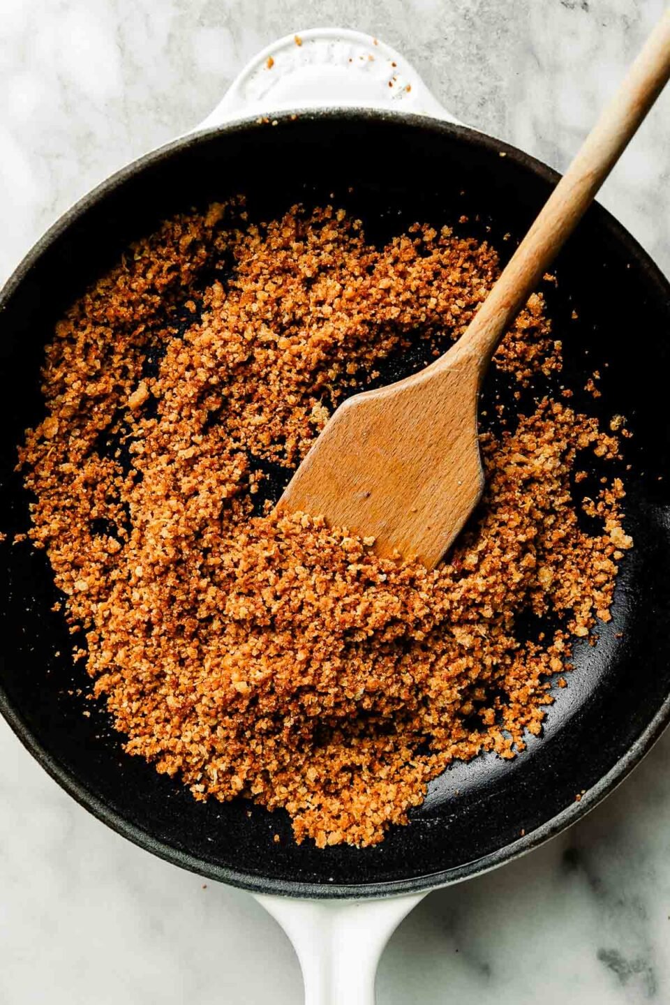 An overhead shot of toasted garlic breadcrumbs with a wooden spoon in a black skillet, sitting atop a white and grey marbled surface.