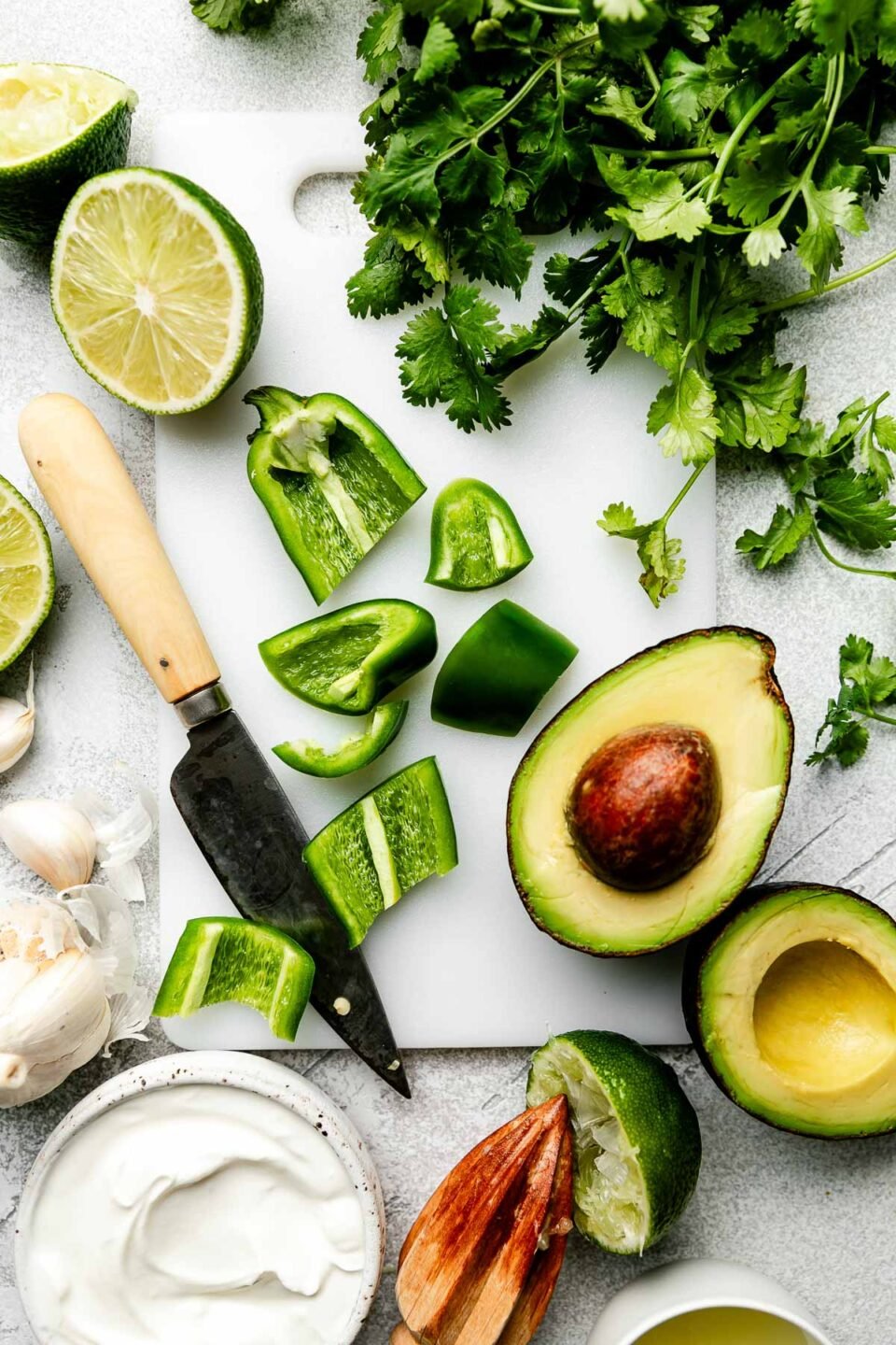 An overhead shot of ingredients displayed on a white cutting board and a grey textured surface: sliced jalapeno, halved avocado, halved limes, sour cream, garlic and cilantro.