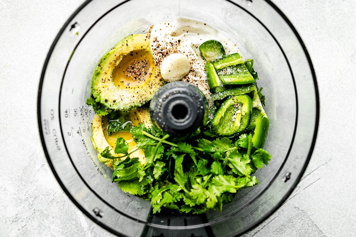 An overhead shot of ingredients in a food processor atop a grey surface: avocado, jalapeno, lime juice, sour cream, garlic, salt and pepper and cilantro.