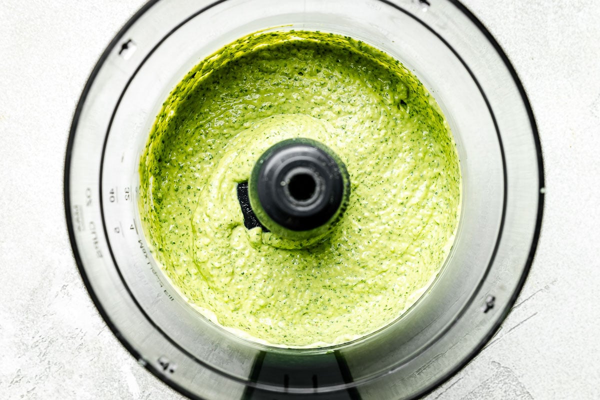 An overhead shot of blended avocado crema in a food processor atop a light grey textured surface.