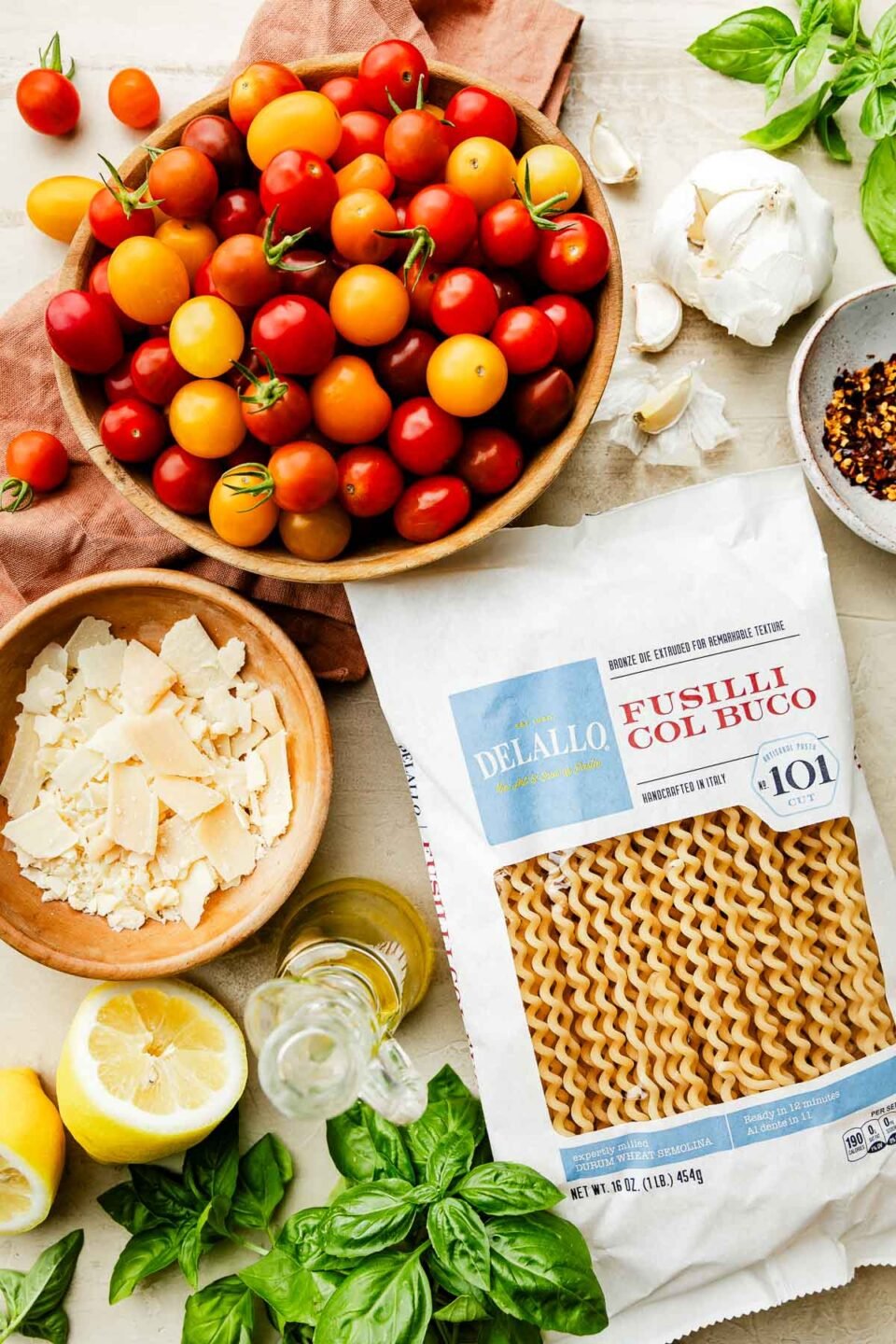 An overhead shot of ingredients displayed on an off-white surface: a bag of fusilli pasta, a bowl of cherry tomatoes, basil, lemon, garlic, red pepper flakes, and shaved parmesan.