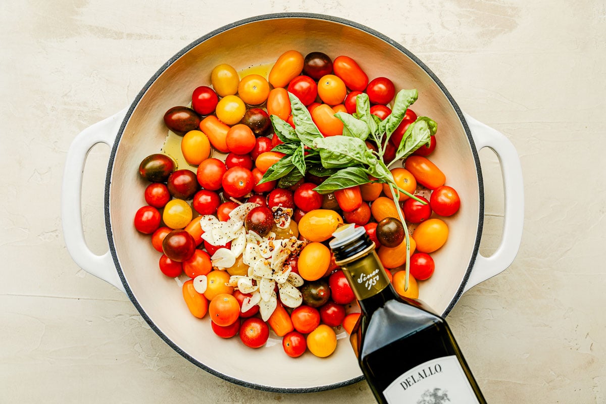 An overhead shot of olive oil being poured onto cherry tomatoes, garlic and basil in a white cast iron skillet.