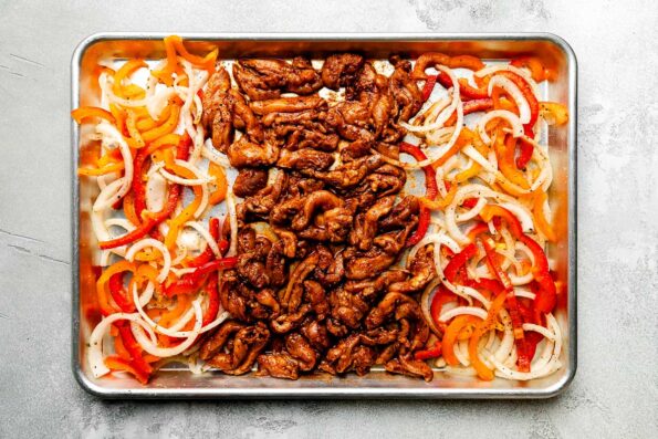 An overhead shot of prepared ingredients on a sheet pan atop a light grey surface: seasoned chicken, sliced bell peppers and onions.