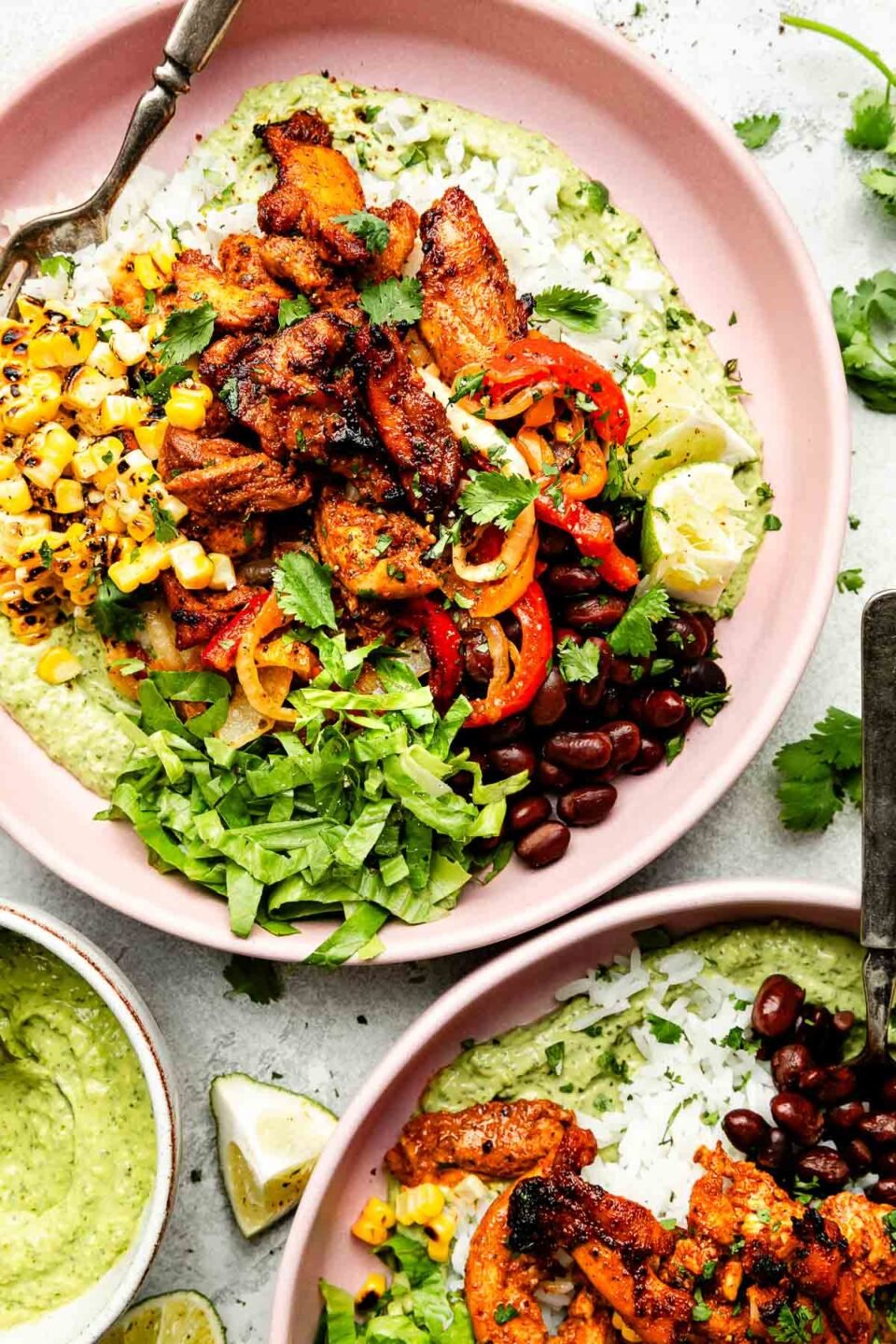 An overhead shot of two assembled fajita bowls on a light pink plate atop a striped cloth on a light grey surface: avocado crema, fajitas, lettuce, corn, pico de gallo, rice and black beans, topped with cilantro. A bowl of crema sits beside the plates.