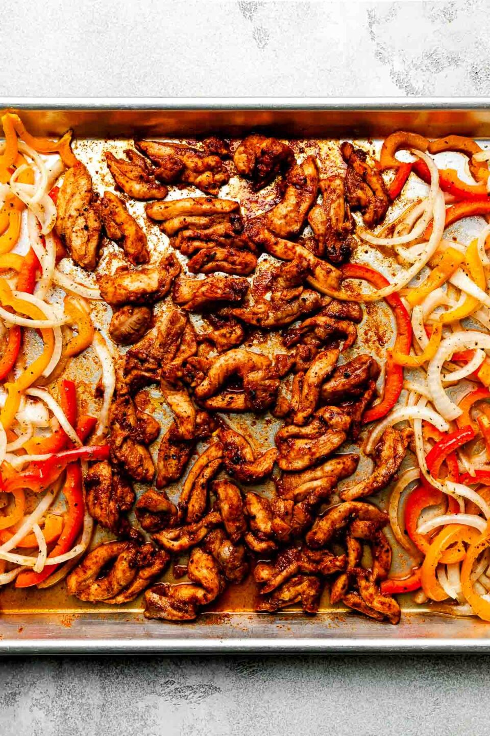 An overhead shot of cooked sheet pan fajitas on a sheet pan atop a light grey surface: seasoned chicken, sliced bell peppers and onions.
