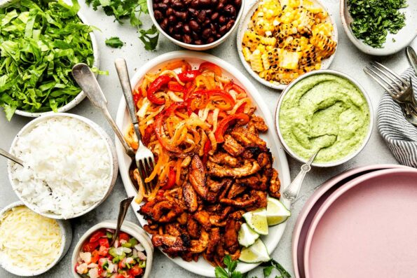 An overhead shot of recipe components displayed in bowls and trays atop a light grey surface: sheet pan chicken fajitas, avocado crema, rice, pico de gallo, charred corn, shredded lettuce, black beans, cheese and chopped cilantro.
