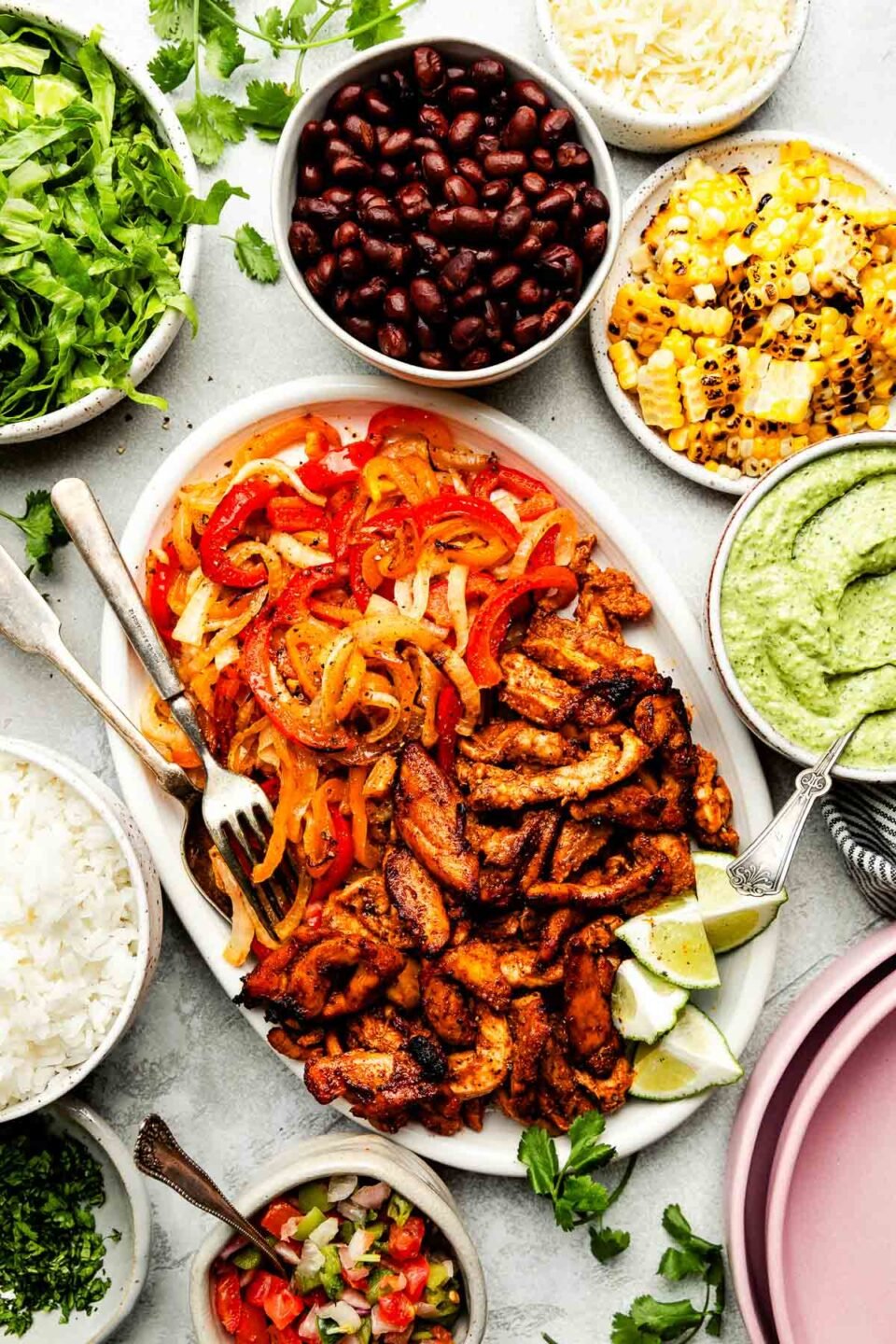 An overhead shot of recipe components displayed in bowls and trays atop a light grey surface: sheet pan chicken fajitas, avocado crema, rice, pico de gallo, charred corn, shredded lettuce, black beans, cheese and chopped cilantro.