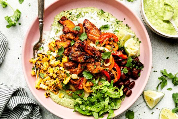 An overhead shot of an assembled fajita bowl on a light pink plate atop a striped cloth on a light grey surface: avocado crema, fajitas, lettuce, corn, pico de gallo, rice and black beans, topped with cilantro.